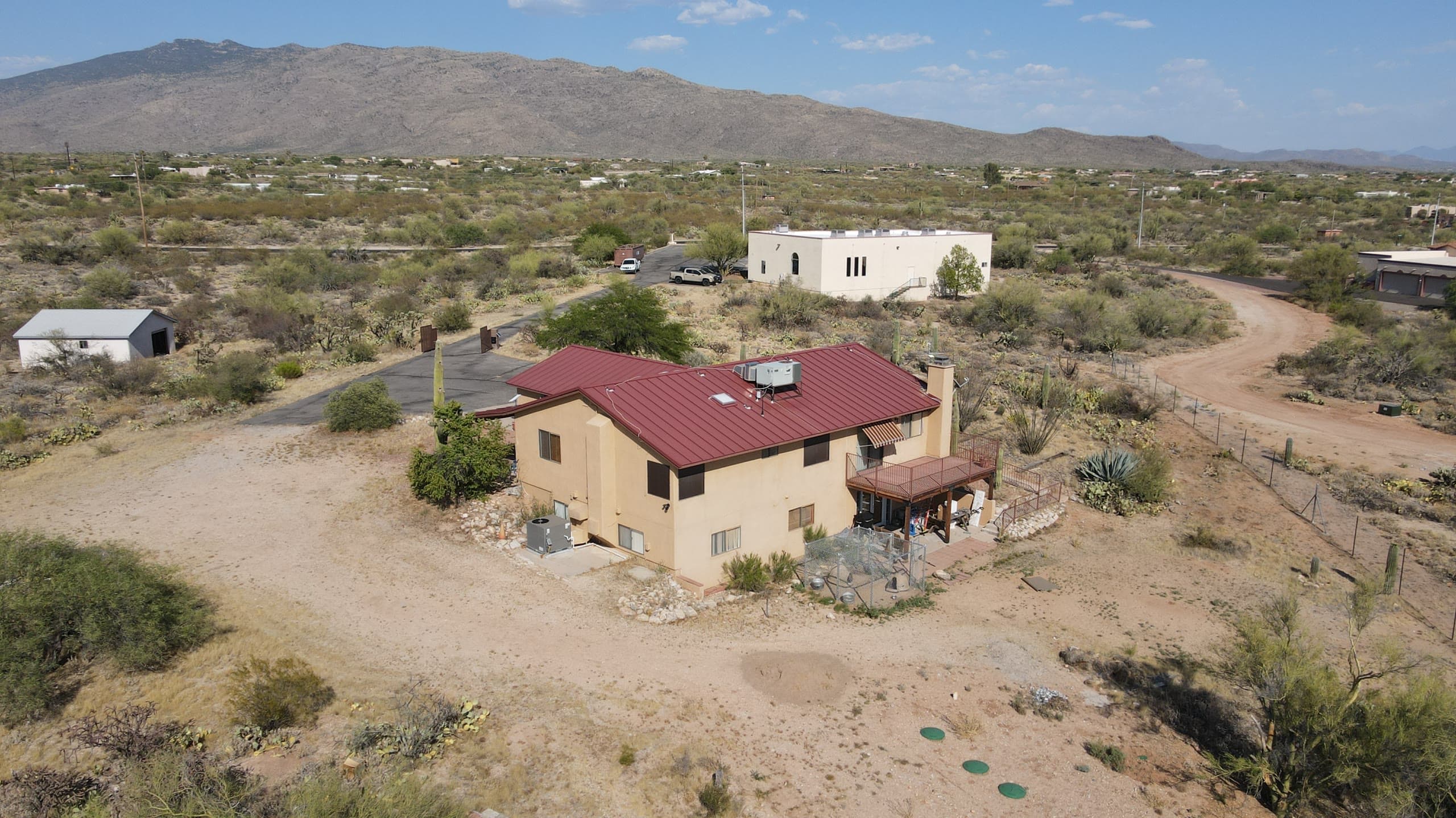 Two-story home with a red metal roof on open acreage, dirt drive and mountains behind