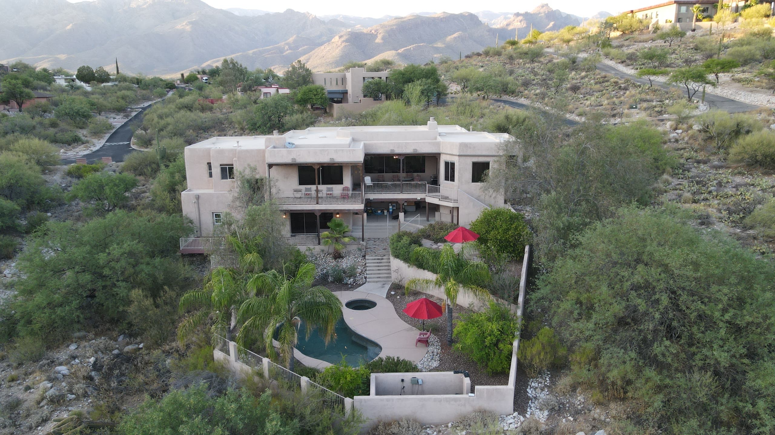 Two-story hillside home with a terraced pool and red umbrellas, mountain ridgelines behind
